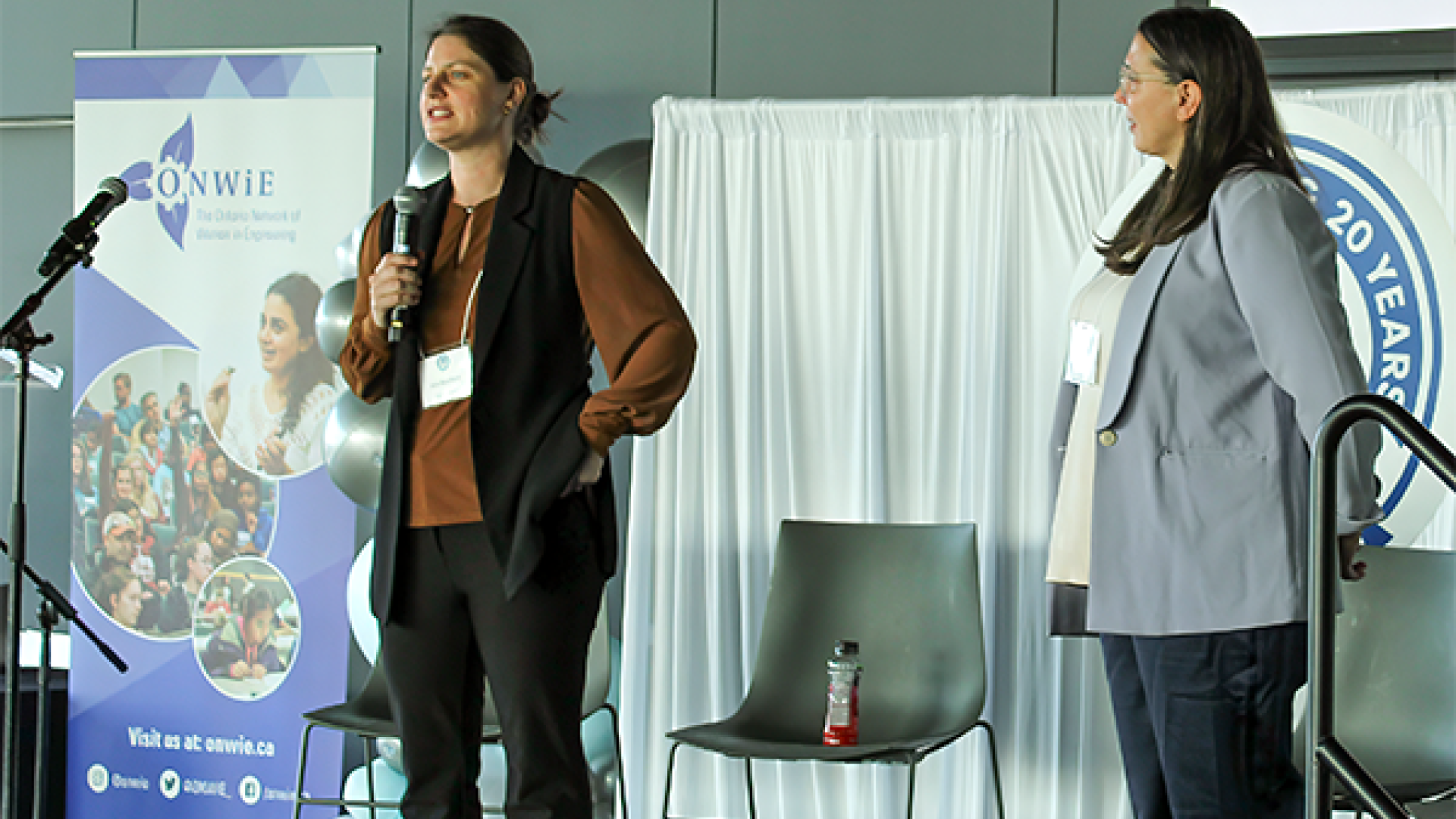 Kim Bouffard and Rebecca White stand on a stage speaking at a conference; one holds a microphone while the other listens. A banner for ONWiE is visible in the background.